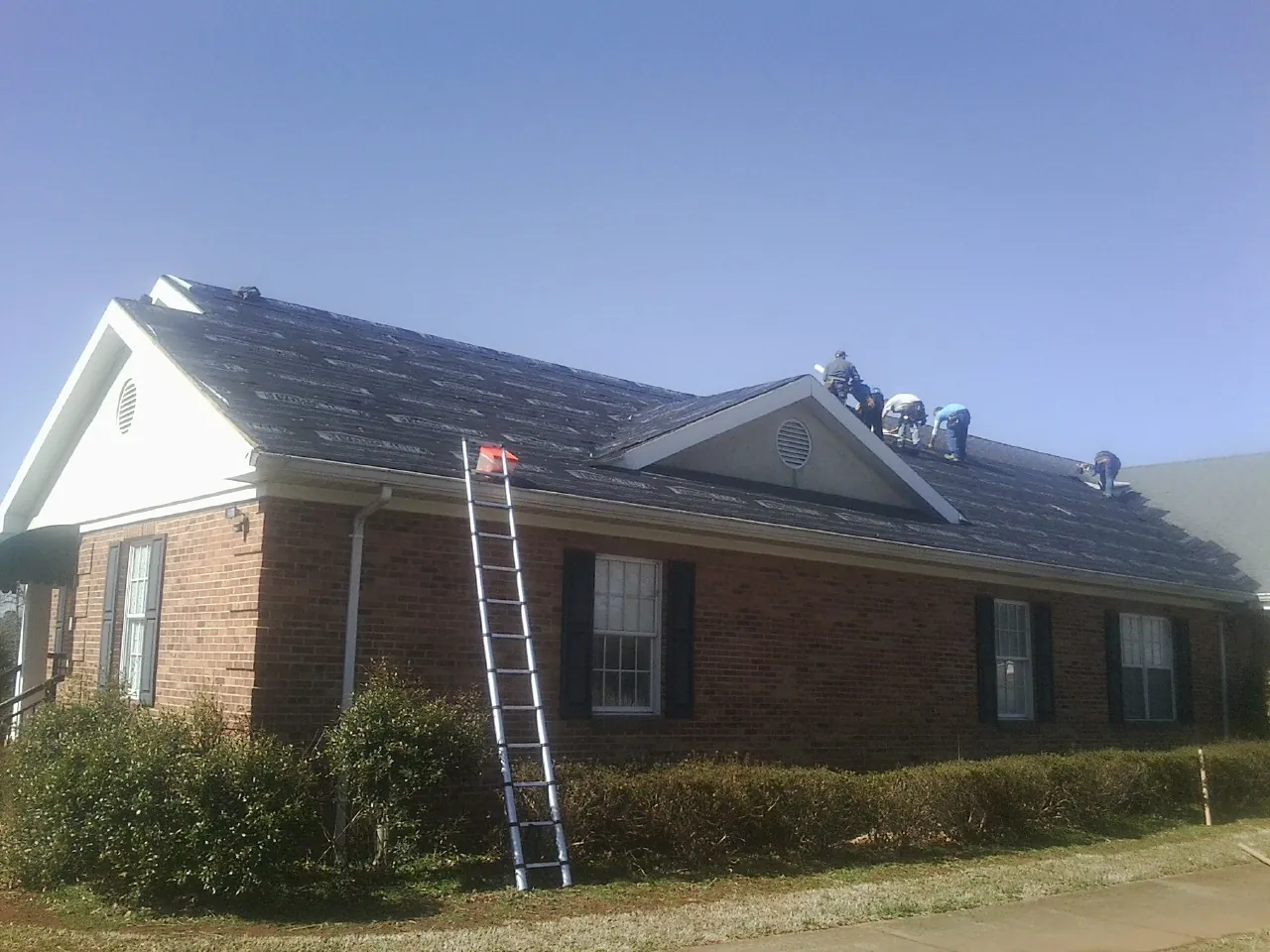 Crew tearing shingles off a brick ranch roof with a ladder set — Emergency Roof Repair work by our crew in Guthrie Center