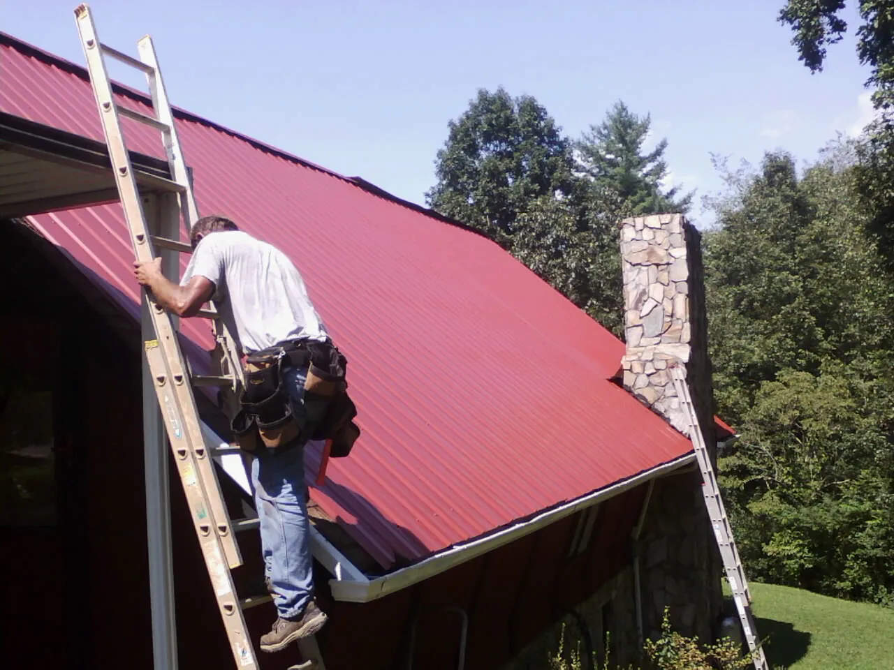 Roofer on a ladder beside a red metal roof and stone chimney — Soffit & Fascia Repair by Roofing Brothers in Guthrie Center